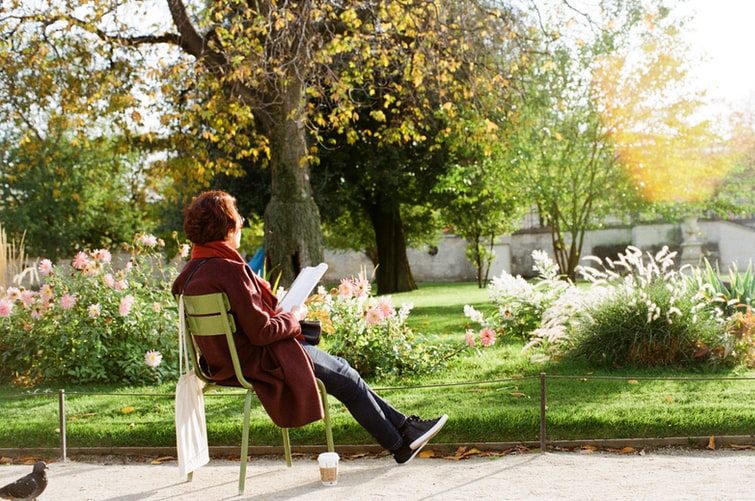 Femeie pe scaun în gradina ei printre flori se relaxeaza si citeste un roman in engleza