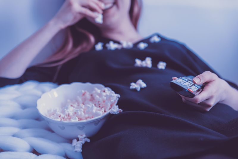 Femeie imbracata in bleumarin urmareste un film in engleza intinsa in pat mancand popcorn dintr-un castron alb cu telecomanda in mana