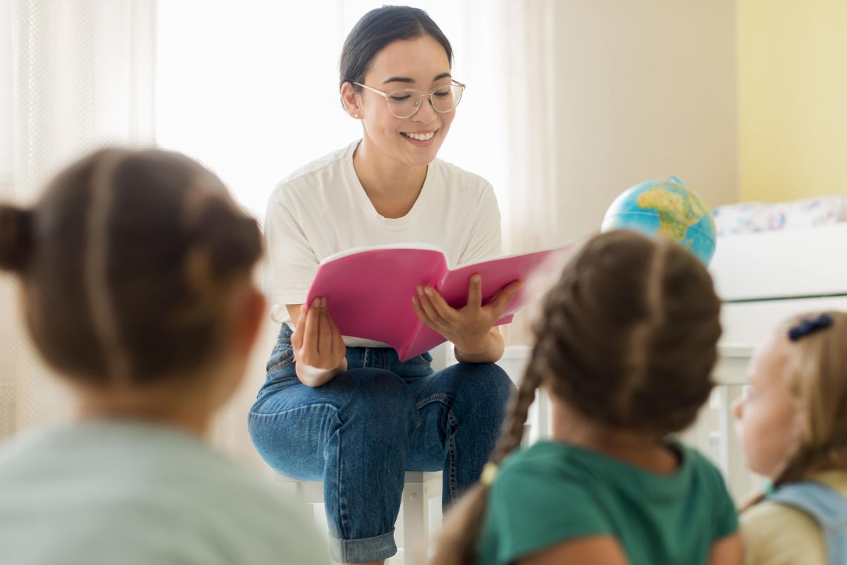profesoara tanara cu chipul bland le citeste copiilor asezati in fata ei, cu spatele la obiectiv, o poveste