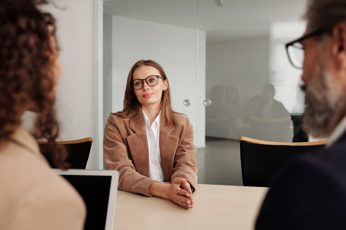 Tanara blonda imbracata office cu ochelari sta la birou in fata a doi recrutori ce ii pun intrebari din CV la un interviu de angajare