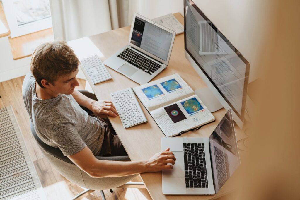 tanar care sta la birou si lucreaza la un desktop si 2 laptopuri simultan pentru a finaliza o sarcina de programare