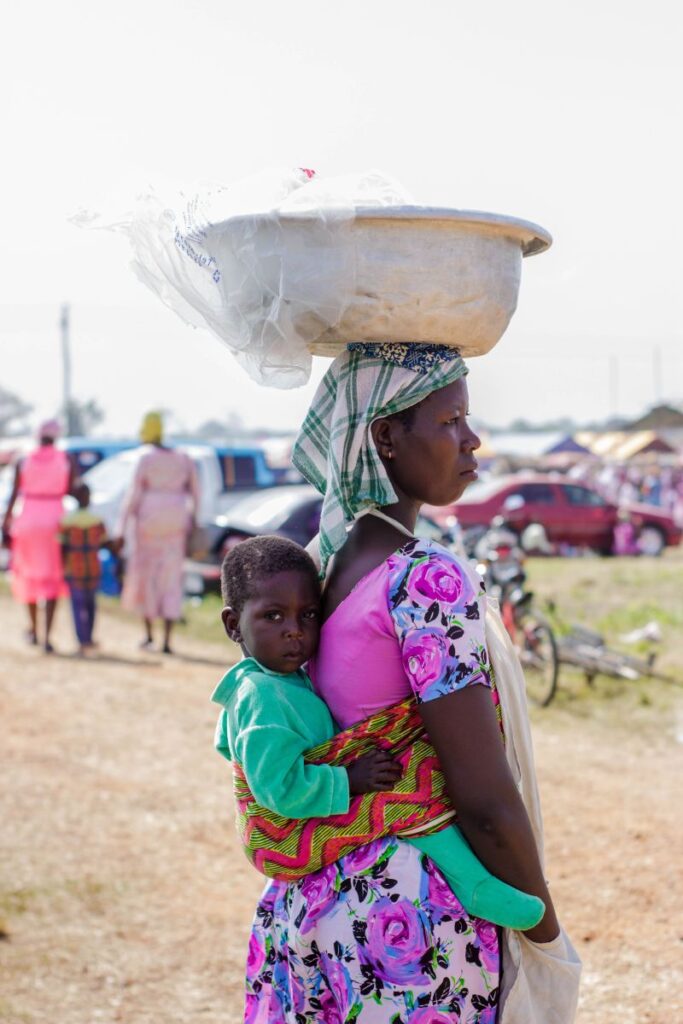 Strada intr-un sat african pe care sta in picioare o mama cu un lighean pe cap si cu copilul ei bebelus purtat in spate