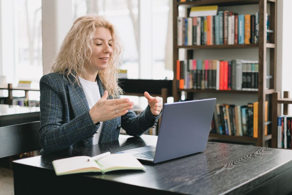 femeie blonda sta la birou in fata unui laptop si invata limba engleza participand la un curs online alaturi de profesor si colegii ei