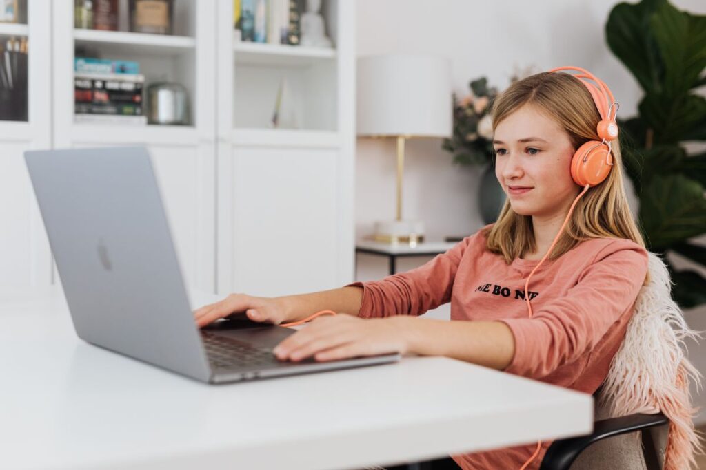 Fata imbracata in portocaliu cu casti la urechi la birou in fata laptopului participa la cursul de engleza online si zambeste incantata