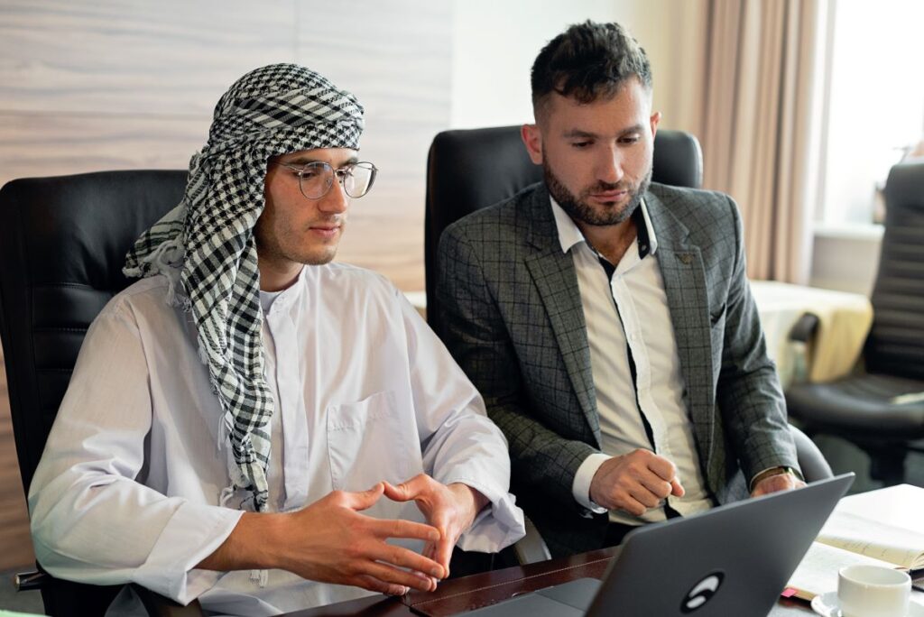 oameni de afaceri european imbracat business si arab cu camasa alba si turban stau alaturi la birou si discuta pe baza cifrelor de pe ecranul unui laptop