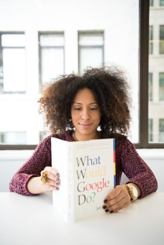 O femeie cu parul mediu cret satena si cu pulover mov si accesorii aurii citeste la o masa alba o carte intitulata What would Google do
