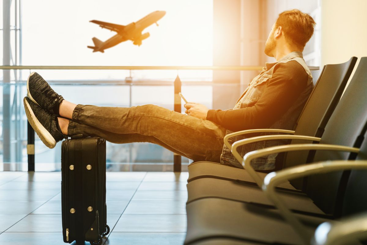 barbat imbracat casual sta singur pe scaun in sala de asteptare la aeroport cu picioarele pe valiza si priveste pe geam un avion care se inalta