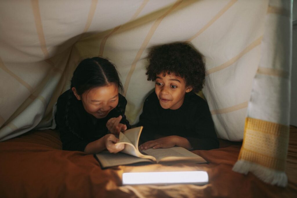 doi copii baiat si fata de varsta scolara mica stau sub plapuma cu o lanterna si rasfoiesc o carte simtindu-se foarte bine