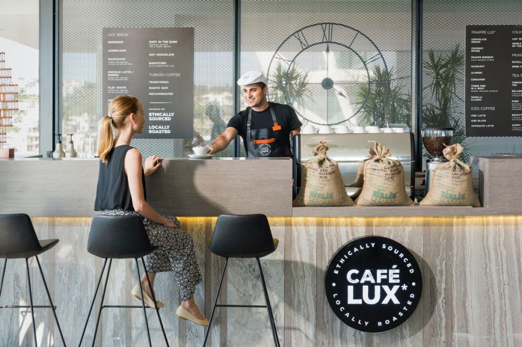 Cafenea in care o clienta blonda sta pe scaun avandu-l in fata ei pe vanzator care ii serveste o cafea pe tejghea, langa niste saci cu boabe de cafea