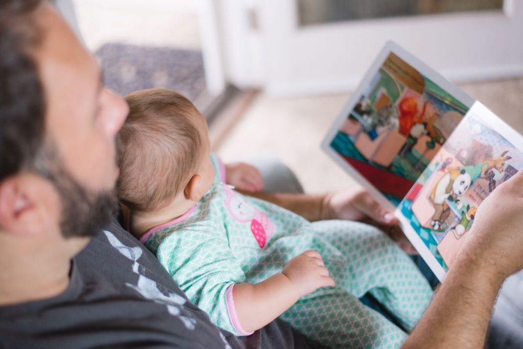 Tata care isi tine bebelusul pe piept si ii citeste o poveste dintr-o carte colorata in engleza