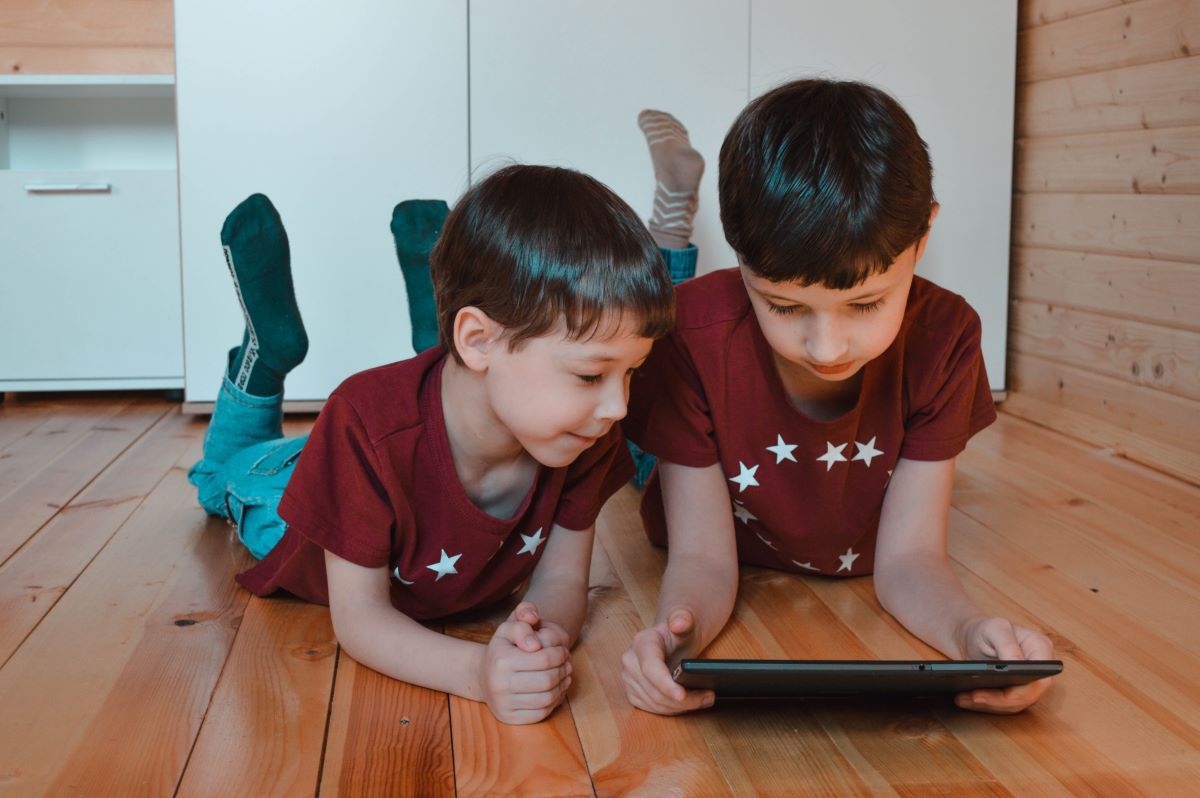 doi baieti prescolari in tricou grena cu stelute si blugi urmaresc la tableta un film in engleza intinsi pe burta pe parchet