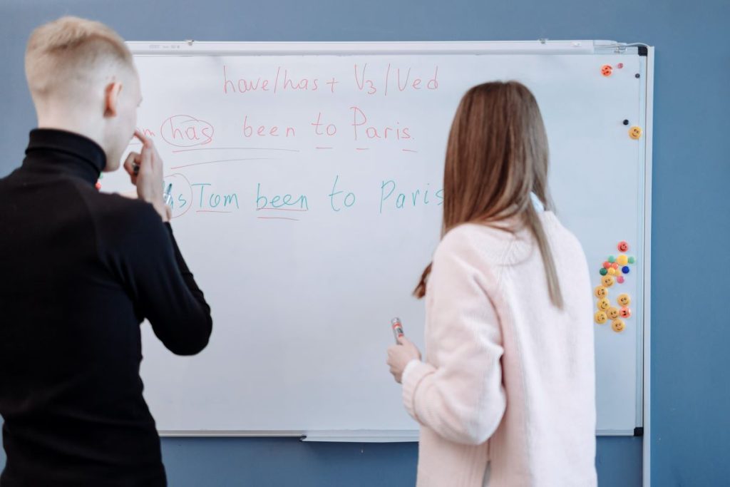 Profesoara si un elev de liceu in sala de clasa la tabla alba stau cu spatele la camera si cu fata la tabla si discuta despre un timp verbal la engleza numit Present Perfect.