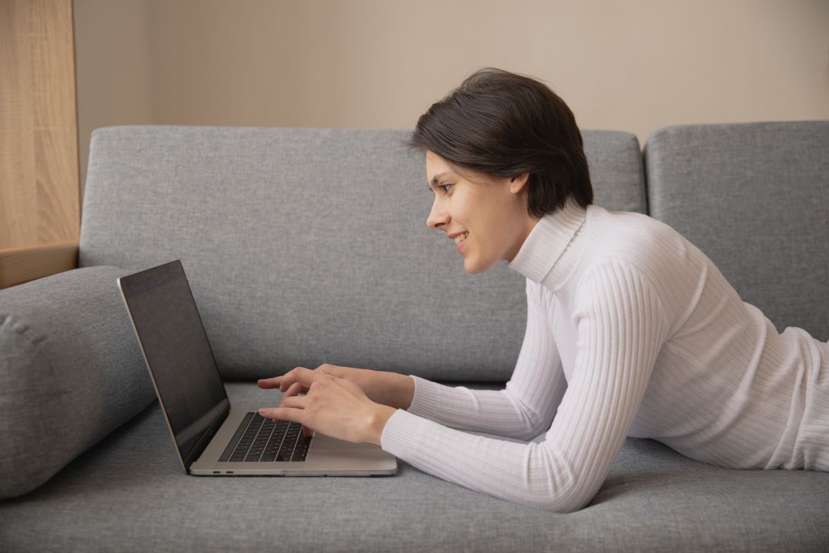Femeie imbracata in alb asezata pe burta pe o canapea gri tasteaza la laptop
