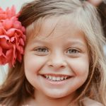 Fetita de 5 ani blonda zambitoare cu o floare mare rosie la ureche