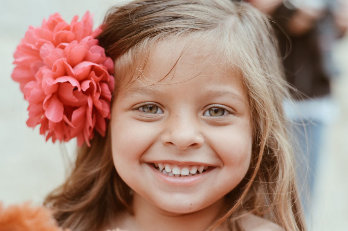 Fetita de 5 ani blonda zambitoare cu o floare mare rosie la ureche