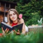 adolescenta sta pe burta pe iarba in fata unei cabane langa un cos de picnic si citeste o carte de dragoste in engleza