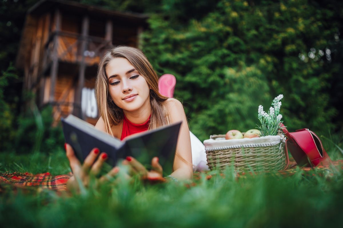 adolescenta sta pe burta pe iarba in fata unei cabane langa un cos de picnic si citeste o carte de dragoste in engleza
