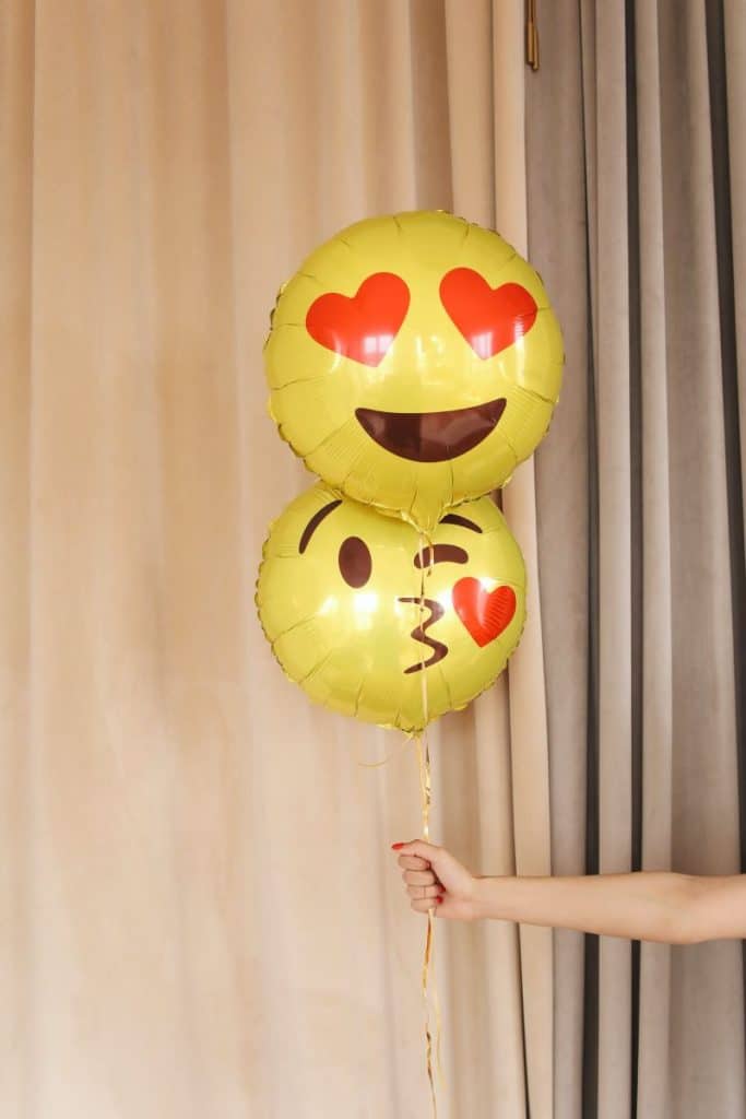 doua baloane cu heliu cu model de emoji galben tinute de o mana de femeie in fata unei draperii crem