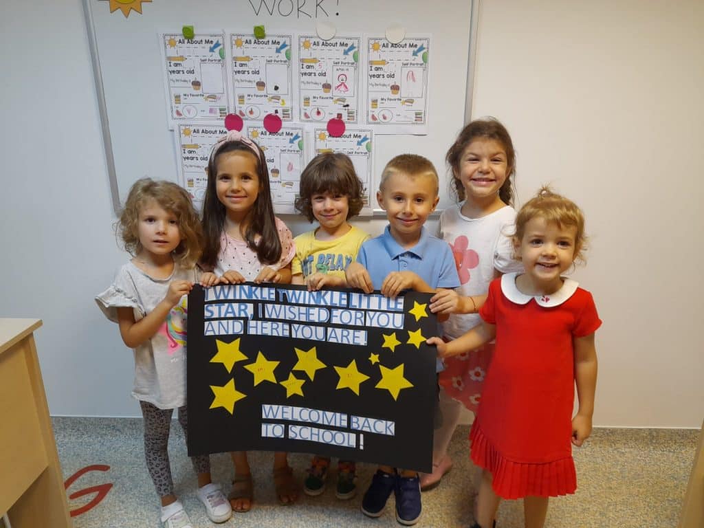 Grup de copii preșcolari zâmbind în fața unui panou decorat cu mesajul Welcome to FollowMe School