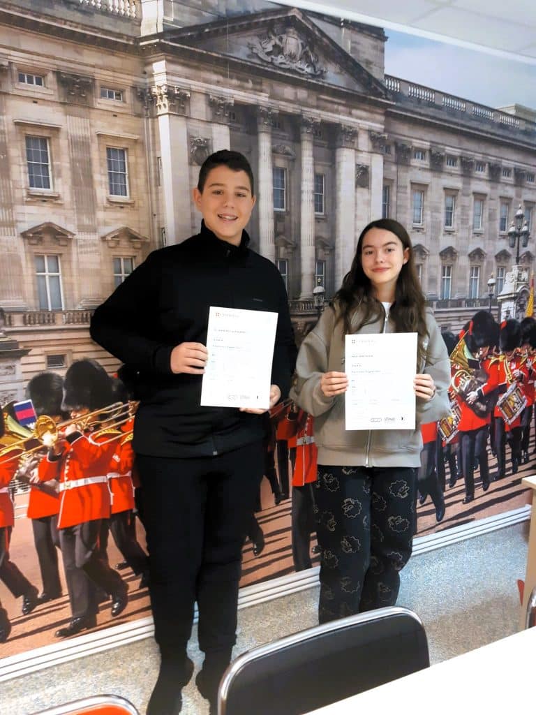 doi adolescenti care au promovat examenul Cambridge arata rezultatele spre camera pe fundalul unui perete cu beefeateri care canta la trompete