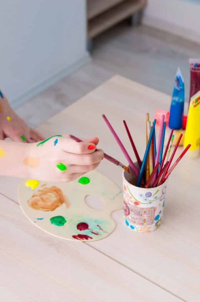 Pahar cu pensule colorate asezat pe masa langa un sevalet cu pete de culoare