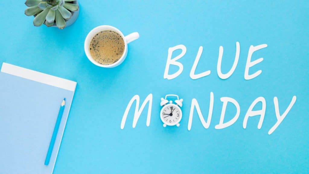 birou cu casti, tastatura si cafea pe fundal albastru peste care scrie blue monday