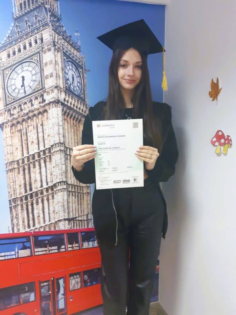 adolescenta bruneta cu parul lung si toga si palarie arata rezultatul la examenul cambridge spre camera pe fundalul unei scene din londra cu Big ben si un doubledecker