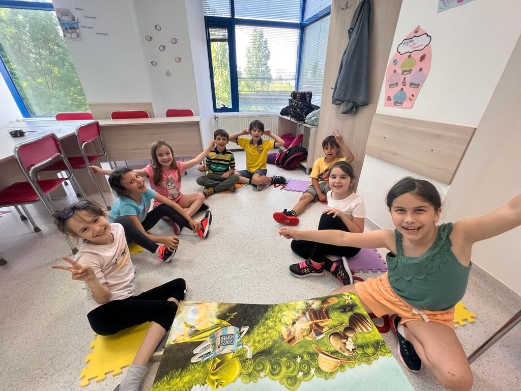 grup de copii care stau pe podea in sala de clasa la cursul de engleza in cerc si zambesc fericiti, avand in fata o carte uriasa de povesti