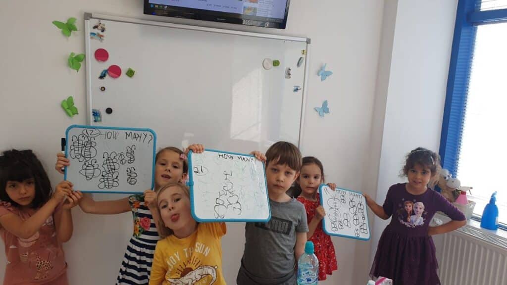 grupe de copii in sala de clasa de la cursul de engleza in picioare in fata tablei arată mini tablitele albe pe care invata să numere si sa exprime pluralul