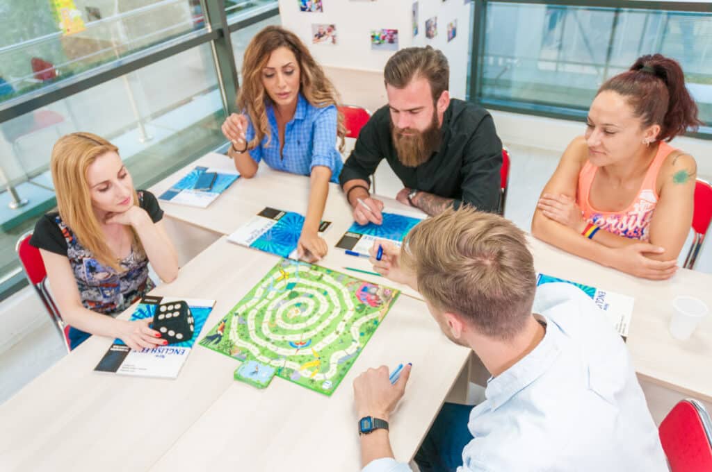 Cursanti tineri in sala de clasa joaca un joc pentru a invata engleza avand in fata manualele pe birou