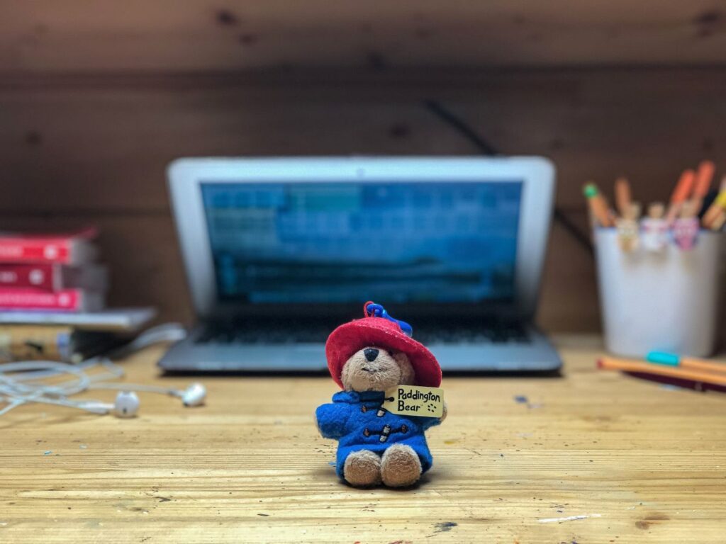 birou de lemn natur pe care se afla un laptop deschis, un teanc de carti, un pahar cu creioane si pixuri blurate si o figurina ursulet Paddington in prim-plan