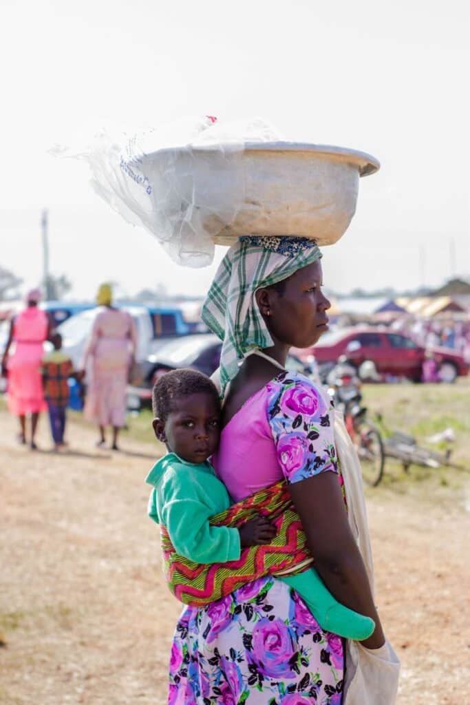 Strada intr-un sat african pe care sta in picioare o mama cu un lighean pe cap si cu copilul ei bebelus purtat in spate