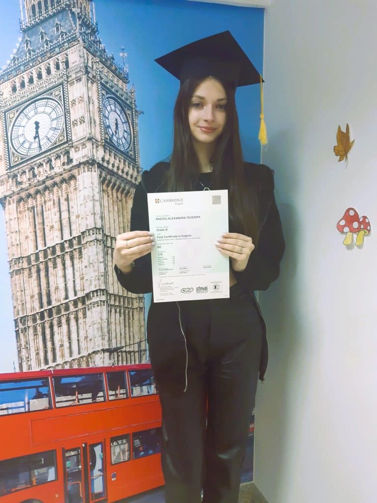 fata cu par lung si palarie pe cap pozeaza in fata unui banner cu simboluri britanice cu diploma obtinuta la examenul cambridge pe care l-a sustinut prin FollowMe