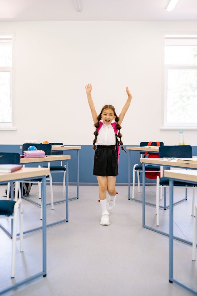 eleva cu parul lung prins in codite in uniforma si ghete albe in sala de clasa ridica mainile in aer si se bucura ca a luat examenul Cambridge