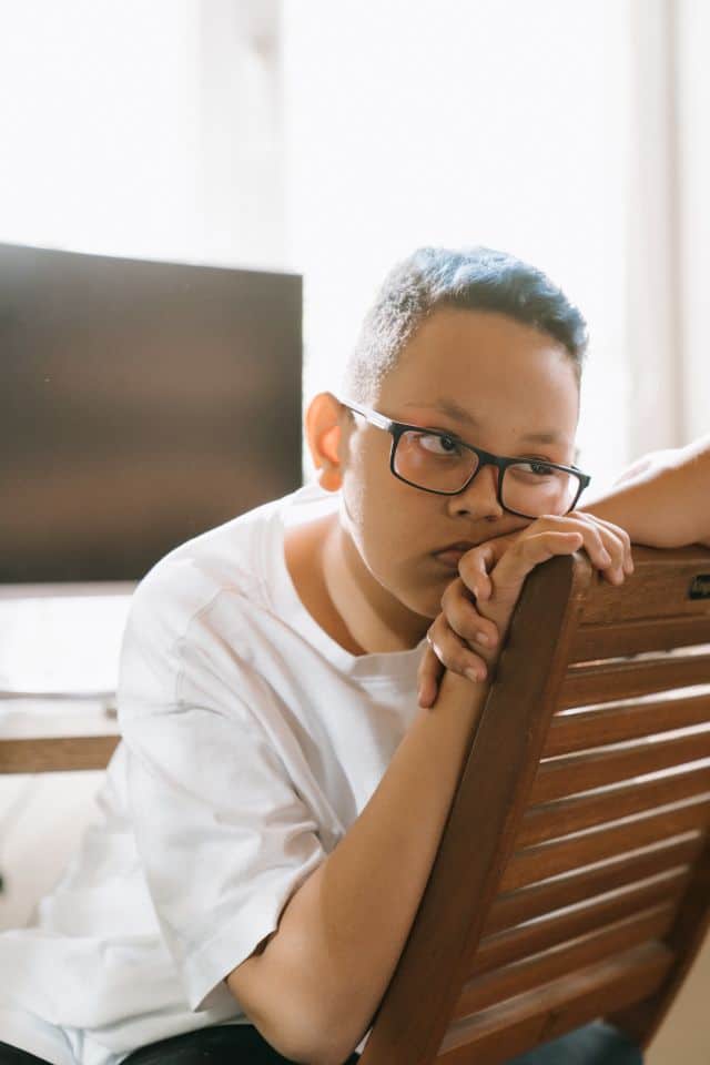 Baiat cu ochelari plictisit isi sprijina barbia pe maini, pe spatarul unui scaun