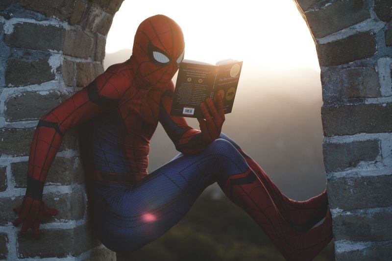 Spiderman asezat in golul de la fereastra unei vechi cladiri citeste o carte avand pe fundal rasaritul soarelui