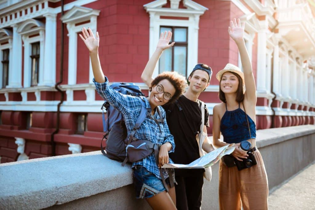 trei tineri imbracati casual stau pe un pod in fata unei cladiri istorice rosii si le fac cu mana altor prieteni pe care si i-au facut in cadrul proiectului Erasmus plus