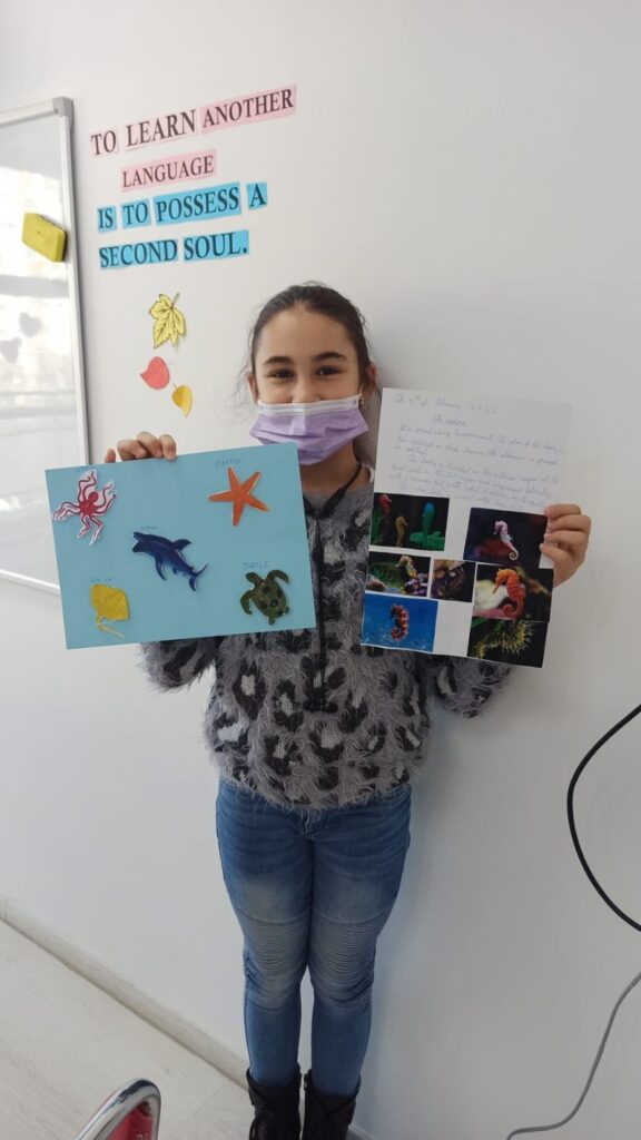 fetita bruneta cu masca ce prezinta la camera proiectele ei cu pesti realizate la cursul de engleza, cu poze si scris