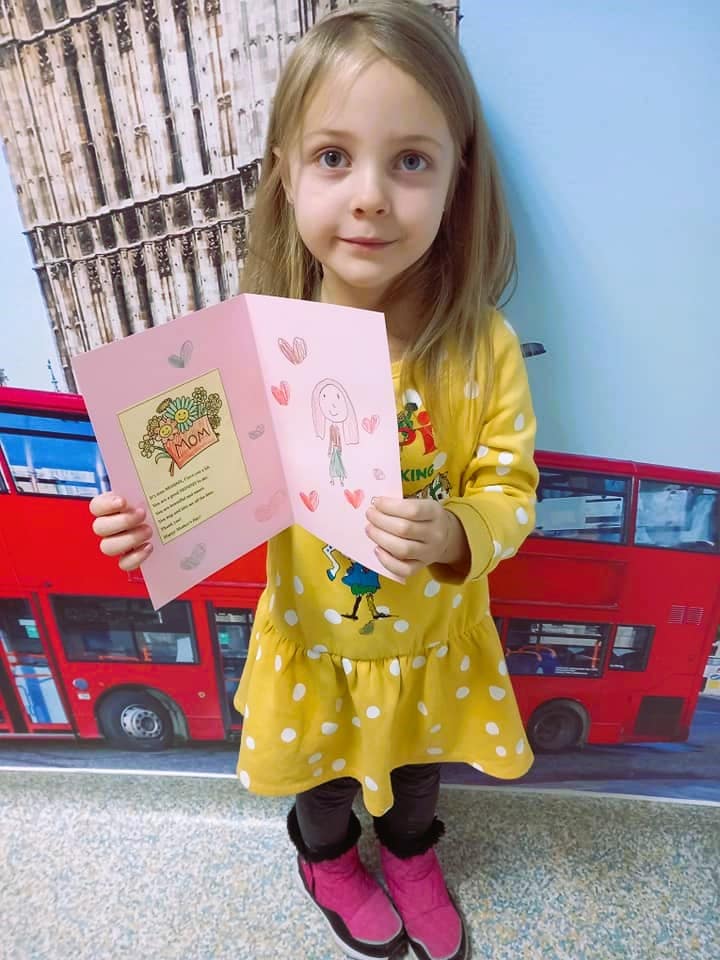 Fetita blonda de 6-7 ani care tine in mana un proiect in engleza care imbina motricitatea cu cunostintele de limba dedicat mamei ei