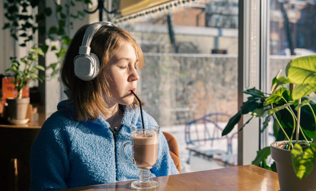 adolescenta roscata imbracata in albastru bea cu paiul cacao cu lapte si asculta la casti povesti in engleza