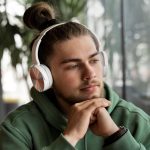 Tanar imbracat in hanorac verde sta cu coatele pe masa si asculta la niste casti mari un podcast sa invete engleza