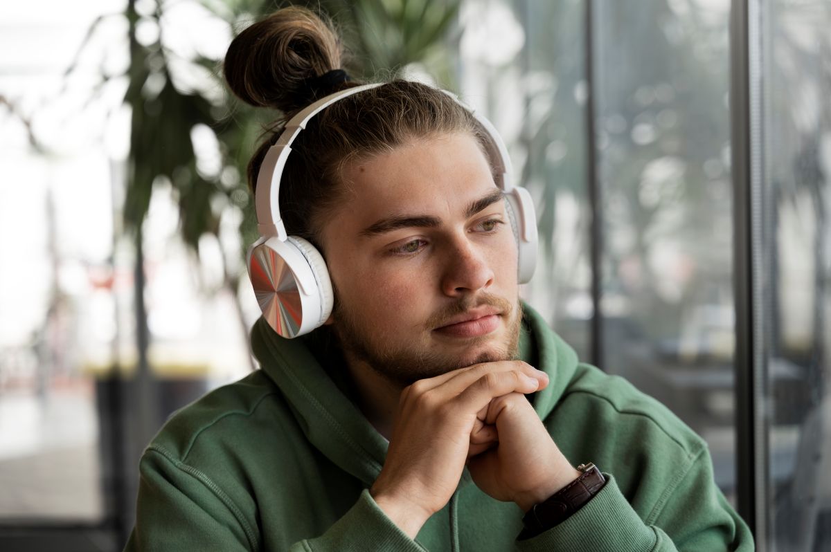 Tanar imbracat in hanorac verde sta cu coatele pe masa si asculta la niste casti mari un podcast sa invete engleza