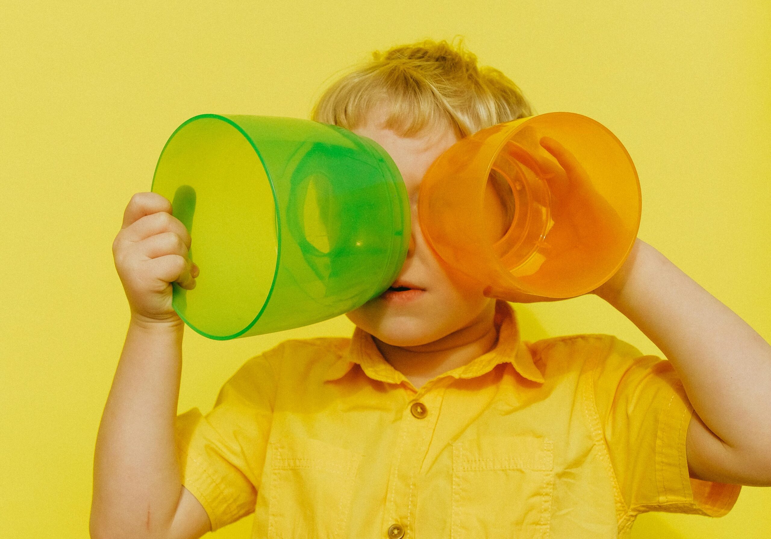 copil in camasa galbena pe fundal galben care tine la ochi doua recipiente de plastic colorate in portocaliu respectiv verde pentru a vedea obiectele din fata lui colorat